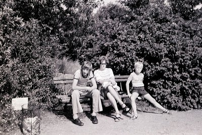 Three children pose on a wooden bench in a lush, green park setting, mid-20th century. The boy on the left wears a striped sh...