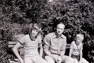 Family portrait in a lush garden setting, mid-20th century. Three individuals seated on a wooden bench surrounded by dense fo...