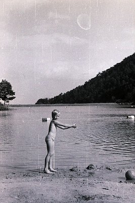 Vintage black-and-white photo of a child in mid-1960s swimwear standing on a rocky lakeside shore, arm extended toward water....