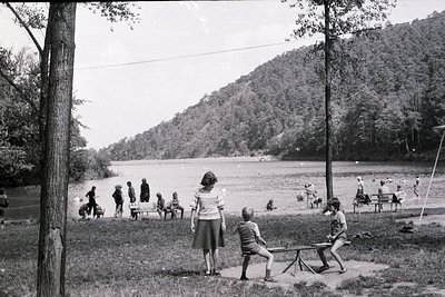 Mid-20th century lakeside gathering with children playing near wooden benches. Lush forested hills frame a serene body of wat...