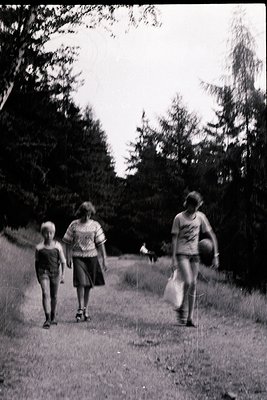 Three figures walk a forested path in mid-20th century attire: woman in patterned dress, child in shorts, adult in T-shirt an...