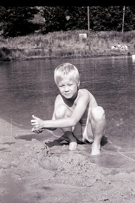 Mid-20th century black-and-white photo of a young boy kneeling in shallow water, holding a small object. Muddy shoreline and ...