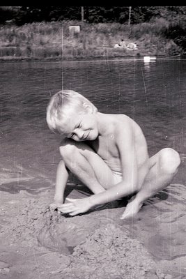 Mid-20th century beach scene: shirtless child kneeling on wet sand, drawing in the tide line. Wooden pier and distant trees f...