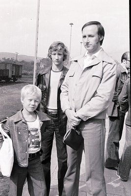 Three individuals pose outdoors near railway tracks in a 1970s-era black-and-white photo. The man wears a light jacket, bell-...