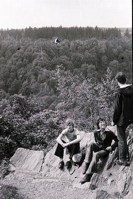 Three individuals pose on a rocky outcrop overlooking dense forest, mid-20th century attire suggests . Lush evergreen trees d...