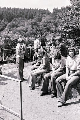 Group of young people in 1970s casual attire—bell-bottoms, long-sleeve shirts, and short hair—sitting/standing by a forested ...