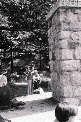 Black-and-white photo of a group of children playing near a stone monument labeled "Frieden" (German for "Peace") in a wooded...
