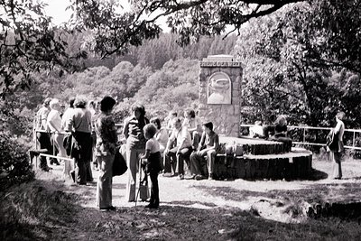 Group gathers around a stone monument with Cyrillic inscription in a forested park, 1960s-70s. Wooden benches and a circular ...