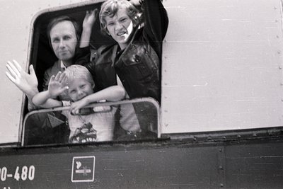 Three individuals wave from a train window, marked "0-480" and featuring a yellow emblem. Mid-20th century European setting, ...