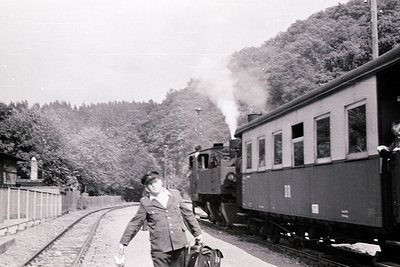 Vintage steam locomotive pulling vintage passenger cars on forested track, mid-20th century. Man in uniform (likely conductor...