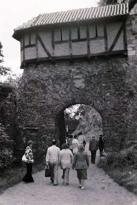 Historic stone archway with timber-framed tower above, likely a medieval fortress entrance. Group of casually dressed visitor...