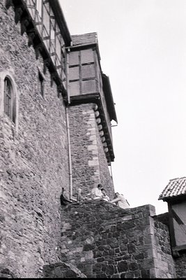 Medieval stone tower with wooden balcony and two figures seated on battlements, likely a fortress or castle. Rustic architect...