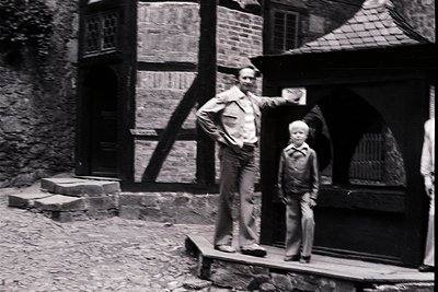 Mid-20th century black-and-white photo of a man and young boy in a European timber-framed courtyard. The man wears a light-co...