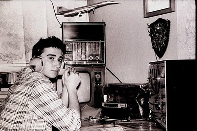Vintage black-and-white photo of a young man in a plaid shirt using a vintage rotary phone in a mid-century living room. Surr...