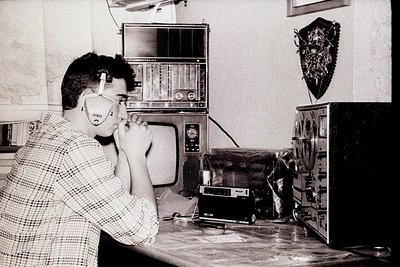Man in 1970s-era home office using vintage corded phone, surrounded by analog audio equipment: reel-to-reel tape recorder, ca...