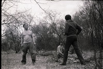 Black-and-white candid of three men in a wooded area, likely 1960s–1970s. One shirtless, one in striped sweater, another in j...