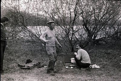 Mid-20th century outdoor scene: shirtless man in sailor-style cap poses by a lakeside campsite. Another man in striped shirt ...