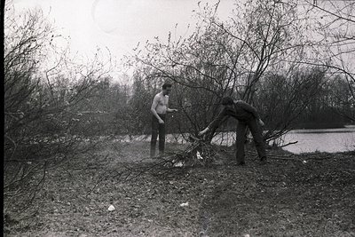 Black-and-white photo of two men clearing fallen branches near a body of water, likely post-storm. One man stands holding a b...