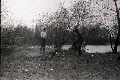 Two men clear fallen branches in a wooded area beside a lake, likely post-storm. Clothing suggests 1960s–1970s era.