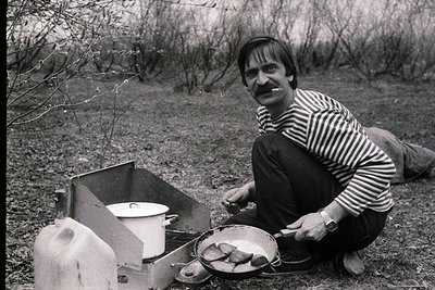 Man in striped sweater cooks fish over portable stove in rural outdoor setting, 1960s–1970s. --- *Notes:* - **Visuals:** Blac...