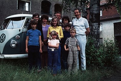 Family portrait in front of a **1970s-era Volkswagen Type 2 (VW Bus) "777"** with residential brick houses in background. Nin...