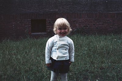 Blond-haired child in a light-colored sweater and striped pants stands in overgrown grass beside a brick wall, mid-20th centu...