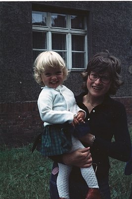 A woman in a dark blouse and glasses holds a laughing toddler in a white sweater, plaid skirt, and tights, set against a bric...