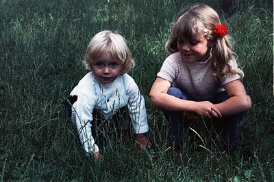 Two young girls in vintage 1970s clothing play in tall grass. The girl on left wears a light-colored, long-sleeved shirt with...