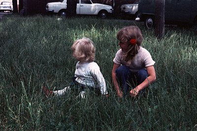 Two children play in tall grass, 1970s suburban setting. Child on left wears striped shirt, child on right sports red bow in ...