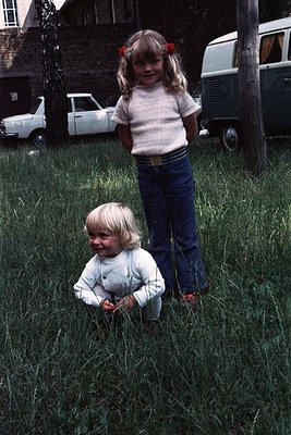 Two children in a residential backyard, 1970s–1980s. Older child (left) wears a striped sweater, blue jeans, and red bows; yo...