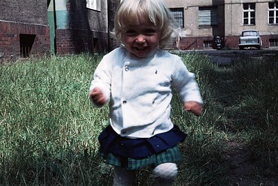 Blond toddler in 1970s-style knit cardigan and plaid skirt running through overgrown grass, mid-laugh. Brick apartment buildi...