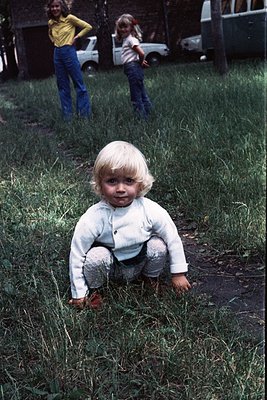 Blond toddler in 1970s-style knitwear kneeling in overgrown grass, mid-outdoor play. Two older children in bell-bottoms and h...