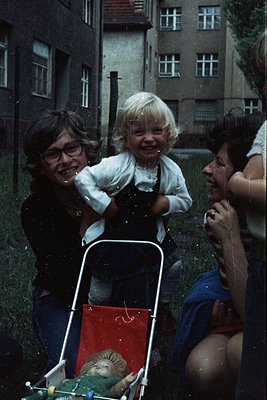 Vintage photo of three adults playfully engaging with a laughing toddler in a red-and-white stroller during a summer rain. Ur...