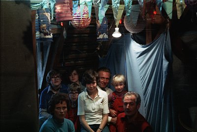 Family portrait in a dimly lit, festive indoor setting, likely a 1970s-1980s home. Seven individuals pose in a semi-circle, f...