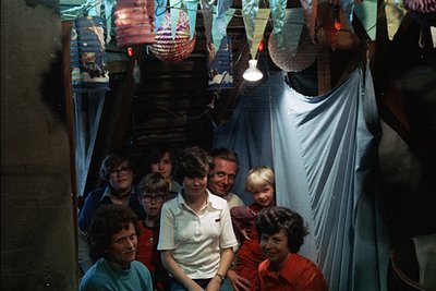 Family portrait in a dimly lit, festive indoor setting, likely a 1970s home. Seven individuals pose in a semi-circle, framed ...