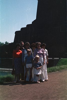 Family portrait from the 1970s, likely Eastern Europe. Seven individuals pose outdoors near a large, angular concrete monumen...