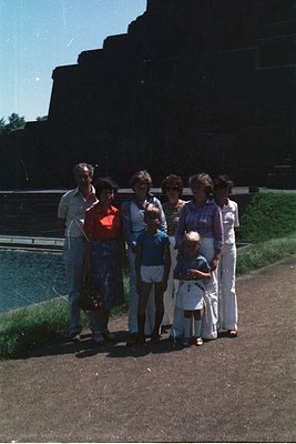 Family portrait by a waterfront structure, likely a dam or reservoir, with concrete steps and greenery. Eight individuals pos...