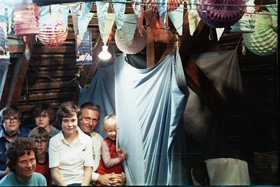 Vintage group portrait inside a rustic, tent-like structure adorned with colorful paper lanterns and triangular flags. Eight ...