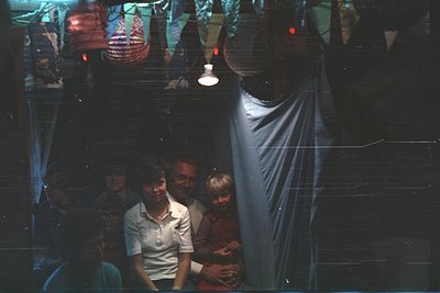 Vintage group portrait in dimly lit indoor setting, likely a 1970s-80s family gathering. Four adults and two children pose cl...