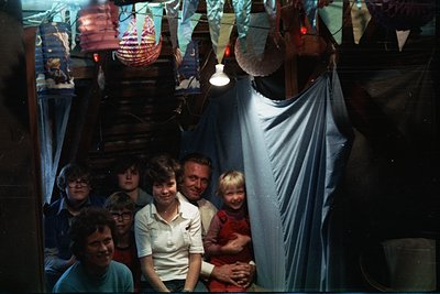 Vintage family portrait in dimly lit indoor setting, likely a 1970s home. Six individuals pose in a semi-circular formation, ...