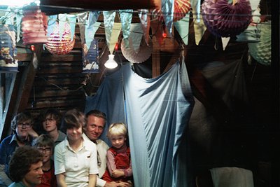 Vintage group photo in a dimly lit, festive setting with paper lanterns and a wooden raftered ceiling. Six adults and a child...