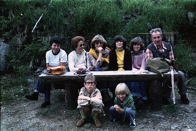 Family group photo on rustic wooden bench in forested area, 1970s. Seven individuals—adults in casual 70s attire (plaid shirt...