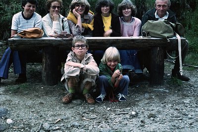 Family portrait on rustic wooden bench in forested setting, 1970s. Adults in striped shirts, cardigans, and casual outerwear;...