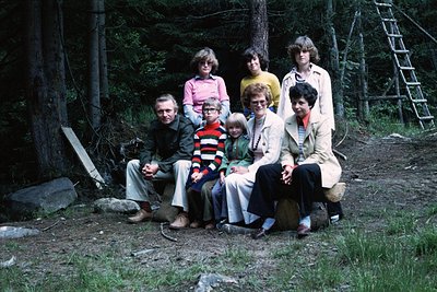 Family group shot in a forested area, likely 1970s. Seven individuals pose on a wooden bench surrounded by trees and a ladder...