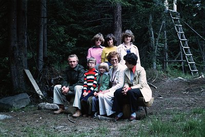 Family group shot in a forested area, 1970s. Seven adults and two children pose on a dirt path surrounded by dense pine trees...