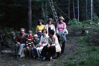 Family group shot in a forested area, likely 1970s. Seven individuals pose on a dirt path surrounded by dense pine trees. Adu...