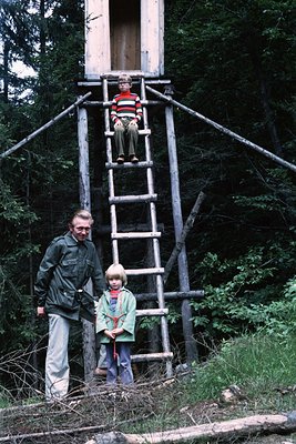 A man in a dark jacket and light pants stands beside a wooden observation tower with a ladder, holding a child’s hand. Anothe...