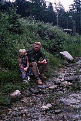 A man and child sit on a rocky forest path, surrounded by dense evergreen foliage. The man wears a short-sleeve shirt, jeans,...
