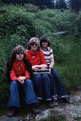 Three individuals pose outdoors in a lush, wooded area, likely a hiking trail, during the 1970s. All wear bell-bottom jeans, ...