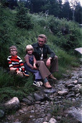 Family portrait in a natural setting, likely mid-20th century (1960s–1970s). Adult wears a long-sleeve striped shirt, dark ja...